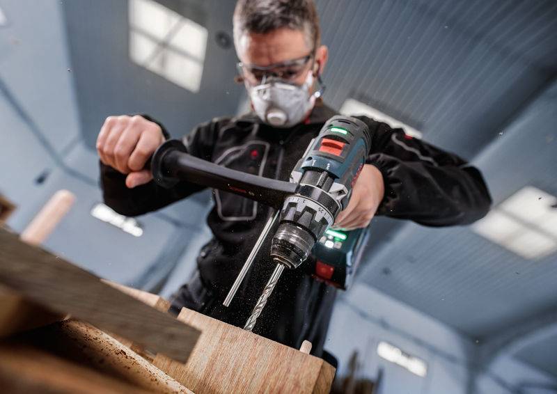 Eine Person, die einen Elektroschrauber auf einer Holzkonstruktion benutzt, trägt Schutzbrille und eine Maske in einer Werkstattatmosphäre.