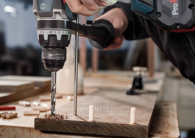 Eine Person benutzt eine Elektrobohrmaschine auf einem Holzbrett und bohrt ein Loch neben Dübeln auf einer Werkbank, was auf ein laufendes Holzbearbeitungsprojekt hindeutet.