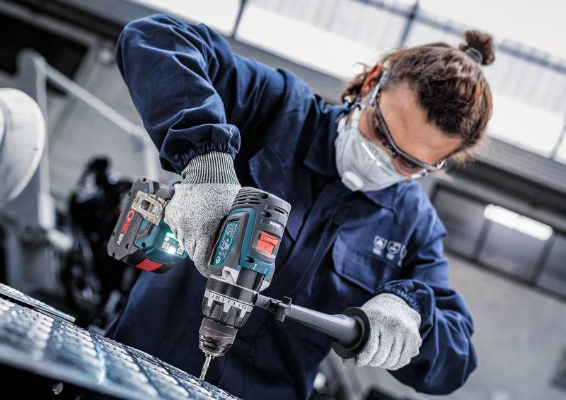 Person in Schutzausrüstung, die eine Bohrmaschine auf Metall in einer Industrieumgebung verwendet.
