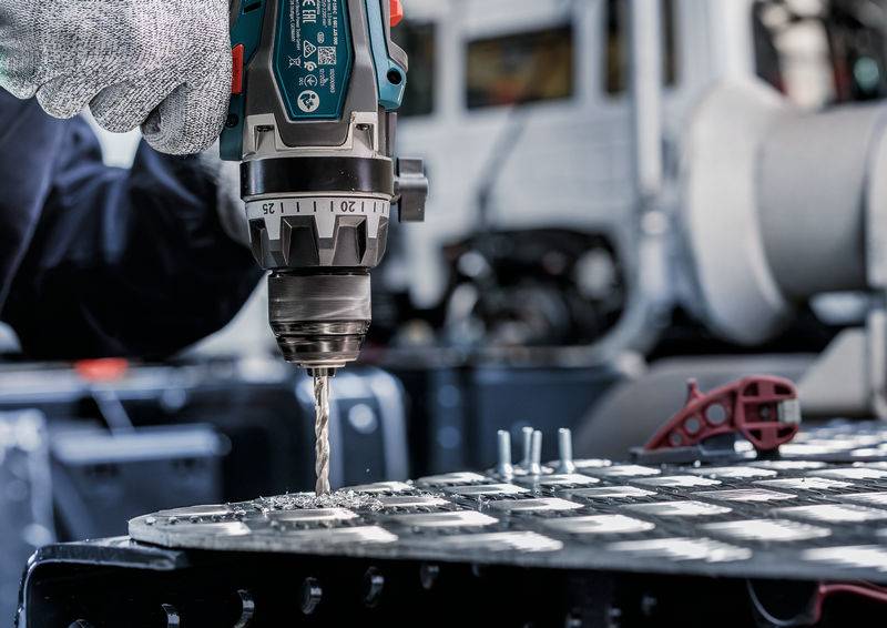 Eine Nahaufnahme einer behandschuhten Hand, die einen Elektroschrauber verwendet, um Löcher in eine Metalloberfläche zu bohren, was Präzision und Industriearbeit veranschaulicht.