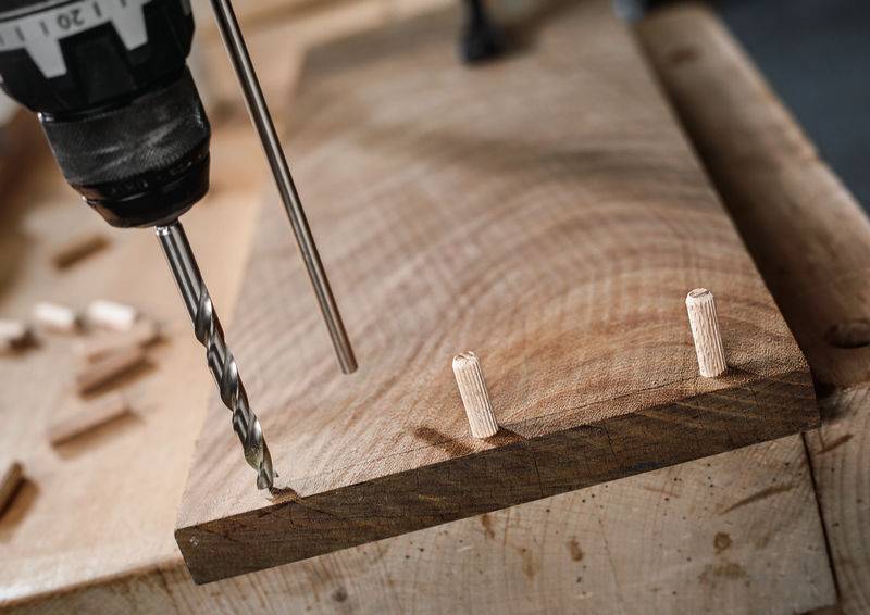 Ein Holzbrett mit zwei eingesetzten Holzdübeln. Ein Elektroschrauber ist positioniert, um ein weiteres Loch zu bohren, was einen Holzbearbeitungsprozess anzeigt.