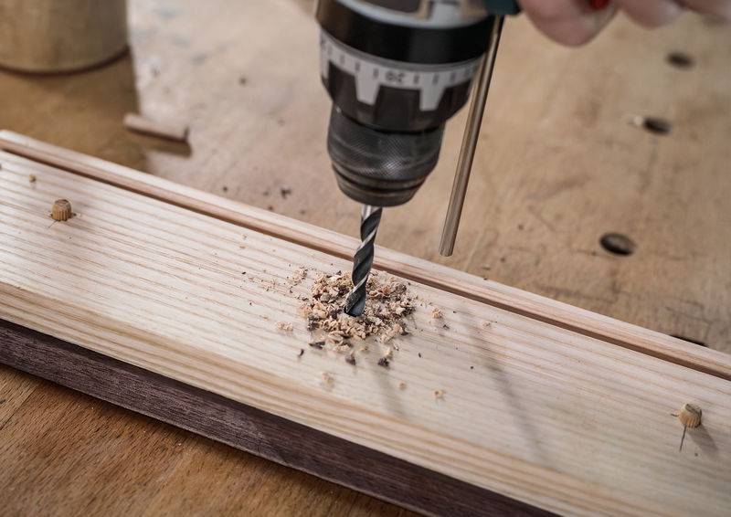 Eine Nahaufnahme einer Bohrmaschine, die ein Loch in ein Holzbrett auf einer Werkbank bohrt, mit Holzspänen, die herum verstreut sind.