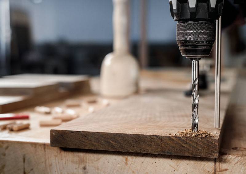 Eine Elektrobohrmaschine mit einem Spiralbohrer bohrt in ein Holzbrett auf einer Werkbank und erzeugt dabei Holzspäne. Werkzeuge und Materialien sind rundherum verstreut.