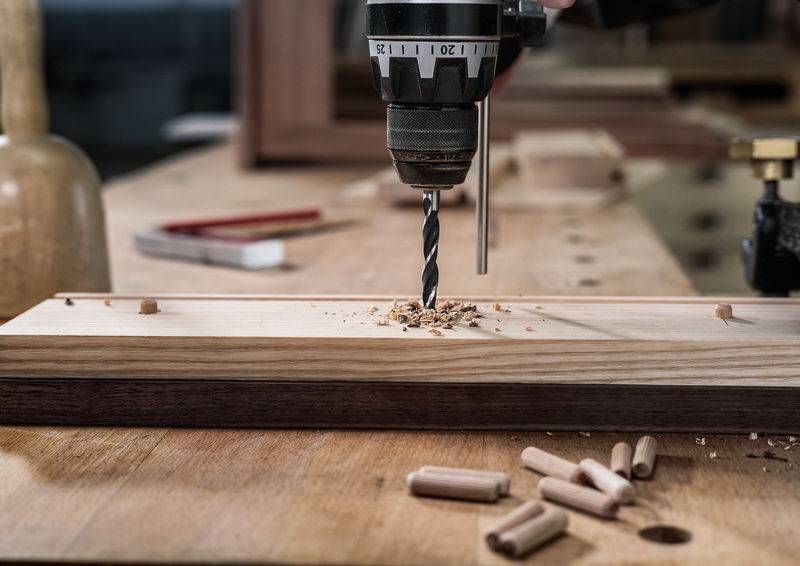 Eine Bohrmaschine bohrt Löcher in ein Holzbrett auf einer Werkbank, umgeben von Sägespänen und Dübeln, was auf laufende Holzarbeiten hindeutet.