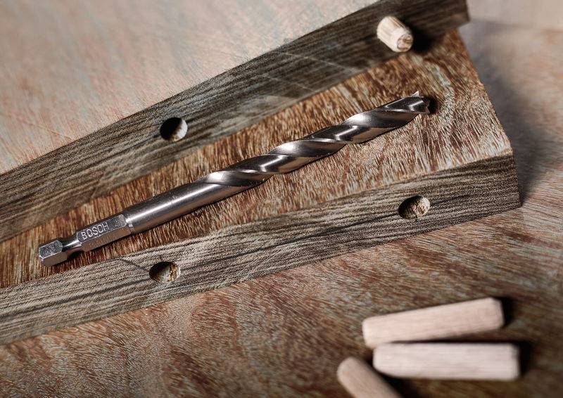 Ein Bohrer wird auf einen Holzblock mit gebohrten Löchern gelegt, zusammen mit hölzernen Stiften, was auf ein Holzbearbeitungs- oder DIY-Projekt hindeutet.