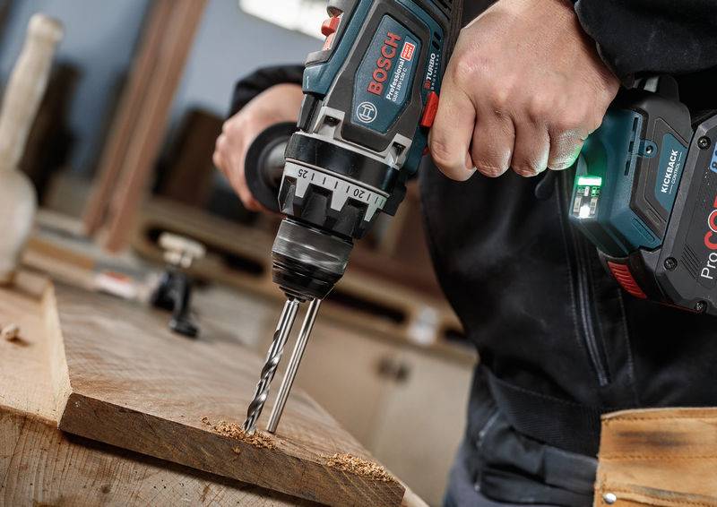 Eine Person benutzt einen Bosch-Akku-Bohrer, um ein Loch in ein Holzstück zu bohren, wobei Holzspäne sichtbar sind und die Verwendung eines Elektrowerkzeugs demonstriert wird.