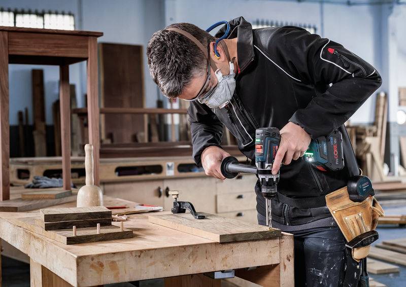 Eine Person in Schutzausrüstung verwendet einen elektrischen Bohrer auf einer Werkbank aus Holz in einer Werkstatt, umgeben von Werkzeugen und Möbelstücken aus Holz.