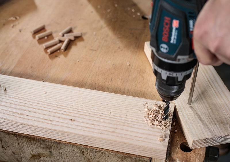 Eine Person benutzt einen Bohrer, um ein Loch in einem Holzbrett auf einer Werkbank zu bohren, wobei Holzdübel und Hobelspäne in der Nähe liegen.