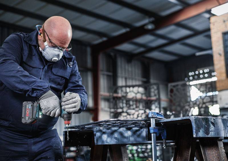 Eine Person in Schutzausrüstung bedient eine Elektrobohrmaschine an einem Holzwerkstück in einer industriellen Werkstattumgebung.
