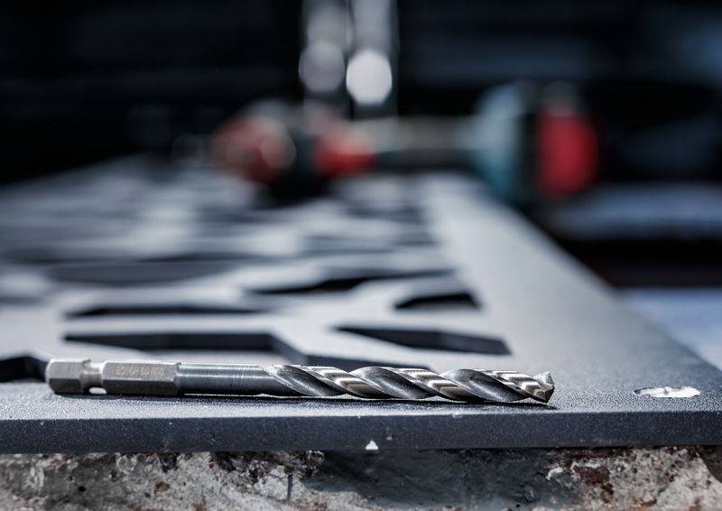 Ein Bohrer ruht auf einer strukturierten, metallischen Oberfläche in einer Werkstattumgebung, mit unscharfen Werkzeugen und Geräten im Hintergrund.