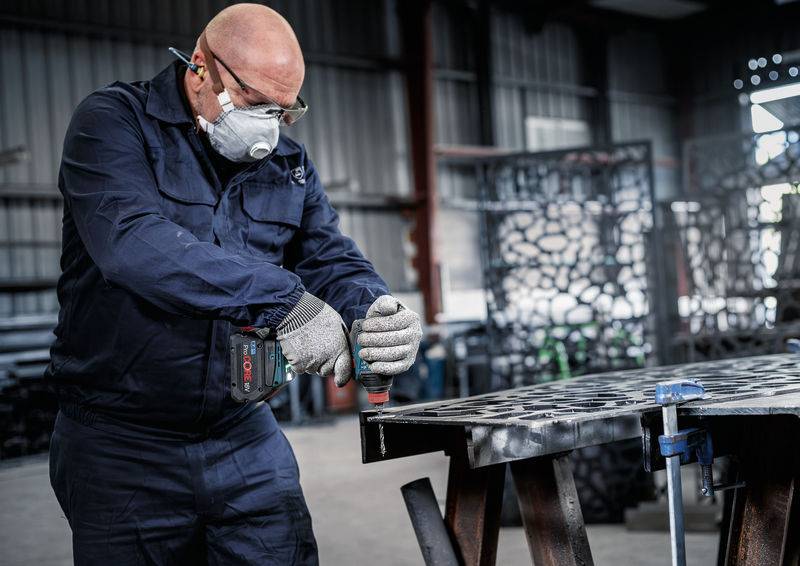Eine Person in Schutzausrüstung bedient ein Elektrowerkzeug auf einer Metalloberfläche in einer industriellen Werkstatt.