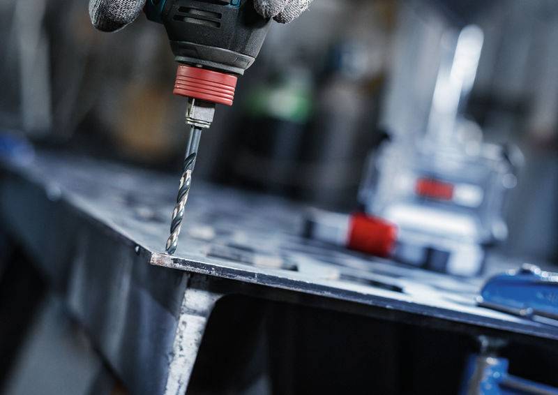 Nahaufnahme einer Elektrobohrmaschine, die Löcher in ein Metallblech bohrt, mit einer behandschuhten Hand, die die Bohrmaschine bedient.