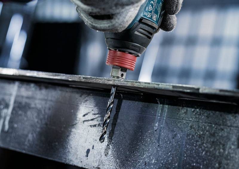 Ein Bohrer mit einem Metallaufsatz bohrt in eine Stahloberfläche ein und verstreut Metallspäne. Die behandschuhte Hand hält den Bohrer sicher an Ort und Stelle.