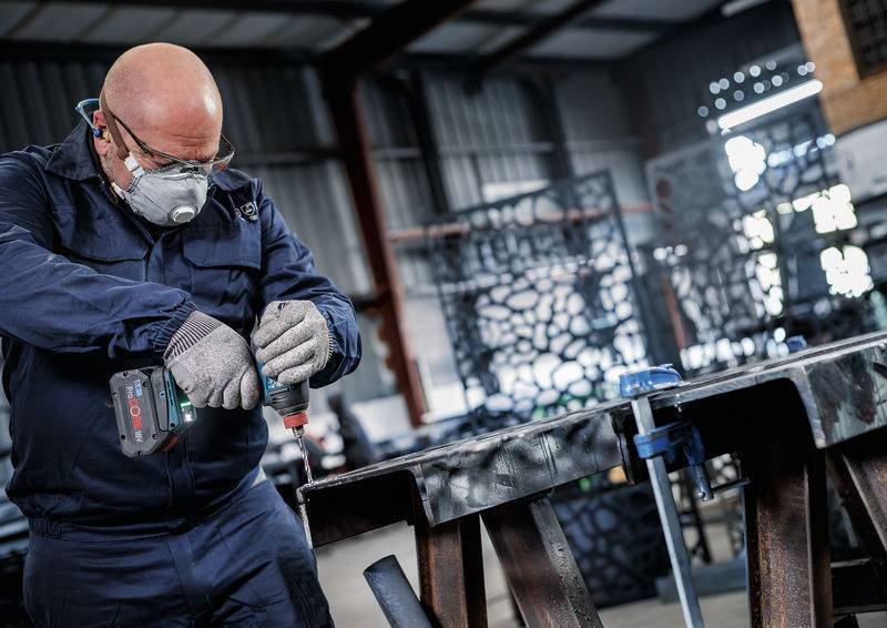 Eine Person, die Schutzausrüstung trägt, benutzt eine Elektrobohrmaschine an einem Metallträger in einer industriellen Werkstattsumgebung.