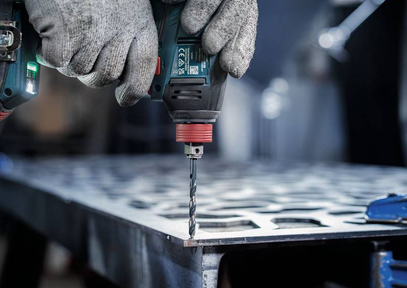 Hände mit Handschuhen benutzen einen Elektroschrauber auf einem Metallblech mit Löchern, was auf eine Metallbearbeitungs- oder Bauaktivität hinweist.