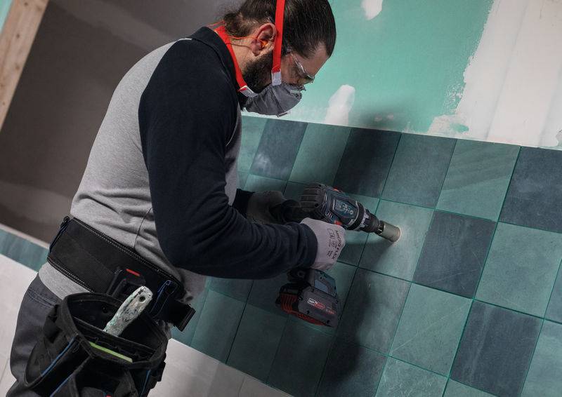 Eine Person, die eine Schutzbrille und Handschuhe trägt, bohrt blaue Fliesen an einer Wand, was auf eine Bau- oder Renovierungstätigkeit hindeutet.