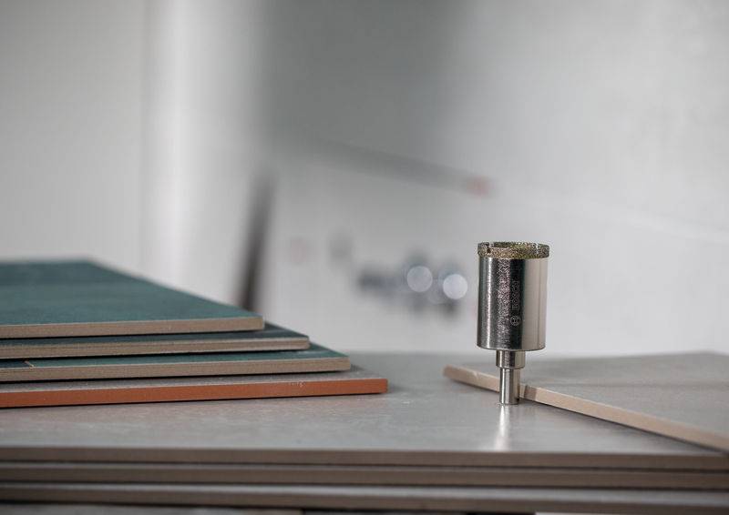 Ein Stapel Holzplatten neben einem Metallbohrer auf einer Werkbank, vor einem unscharfen Hintergrund, was auf eine Holzbearbeitungsszene hindeutet.