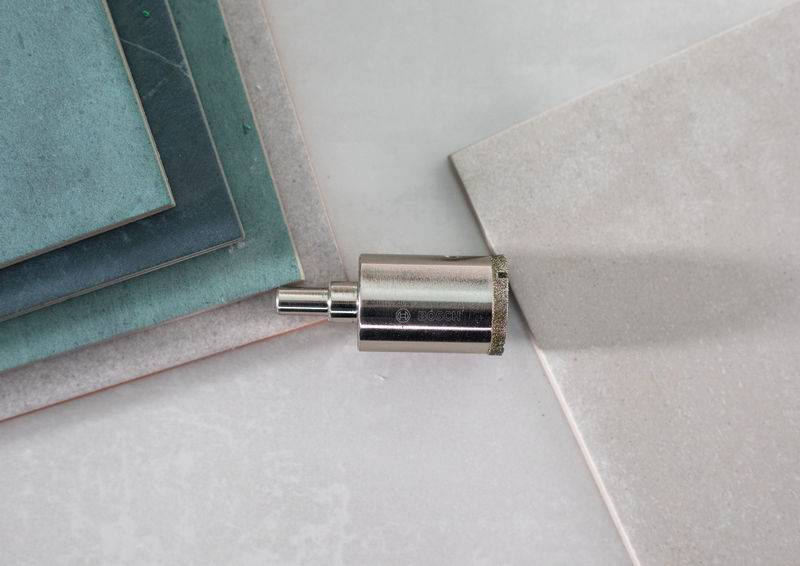 Mehrere Fliesen in unterschiedlichen Blau- und Grautönen sind neben einem silbernen Bohraufsatz auf einer hellgrauen Oberfläche angeordnet.