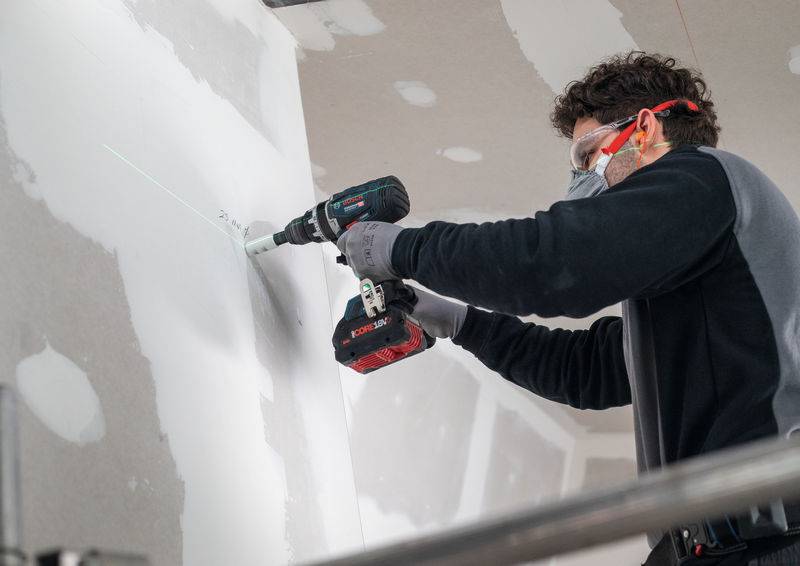 Person trägt Schutzausrüstung und benutzt einen Elektrobohrer an einer weißen Wand, wobei der Fokus auf Bau- oder Renovierungsarbeiten liegt.