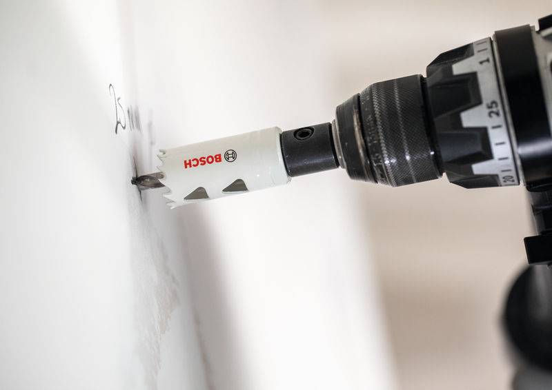 Ein Elektroboherer mit Lochsägenaufsatz bohrt in eine weiße Wand und erzeugt dabei feinen Staub um die Eintrittsstelle.