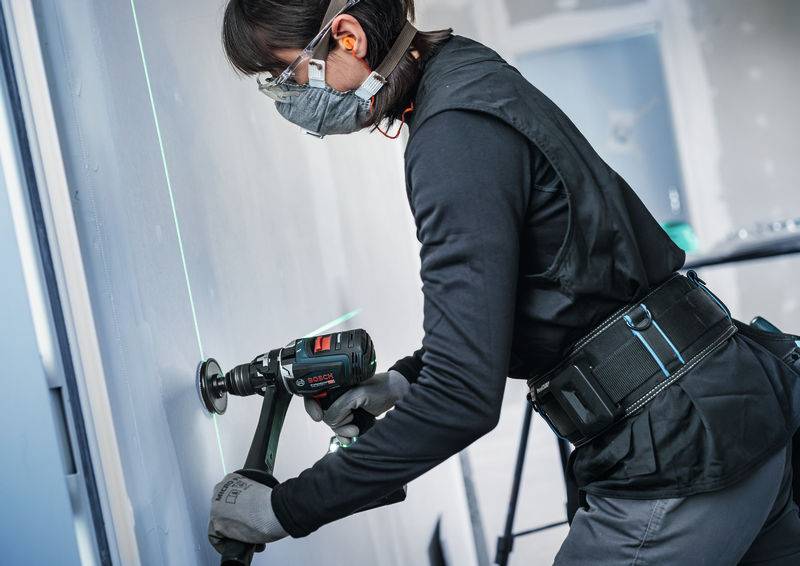 Ein Bauarbeiter, der Schutzausrüstung trägt, nutzt einen Bohrer, um ein Loch in eine Wand zu bohren, dabei wird er durch eine grüne Laserlinie zur Präzision angeleitet.