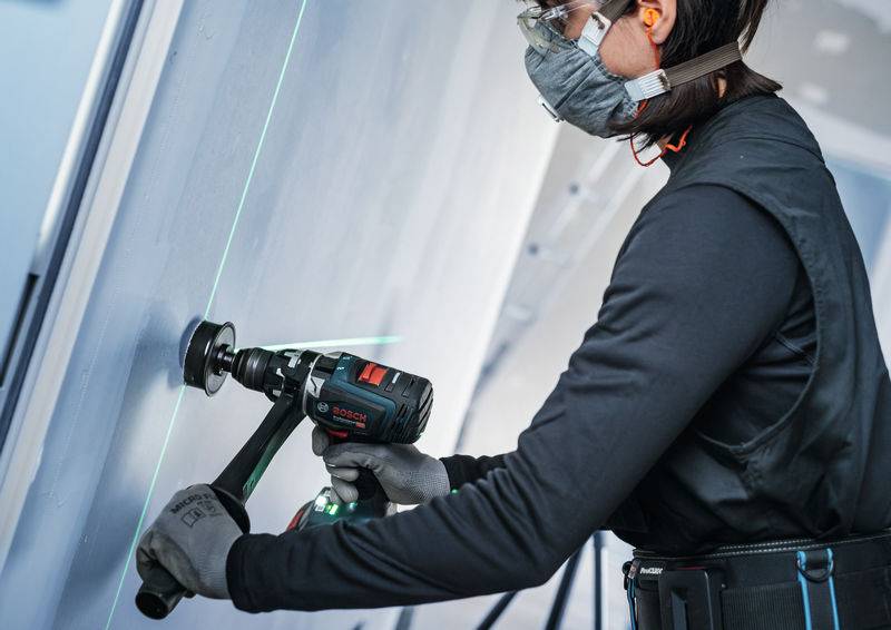 Eine Person in Schutzausrüstung verwendet ein Bosch-Elektrowerkzeug, um mit Hilfe einer grünen Laserlinie für Präzision in ein großes Brett zu bohren.