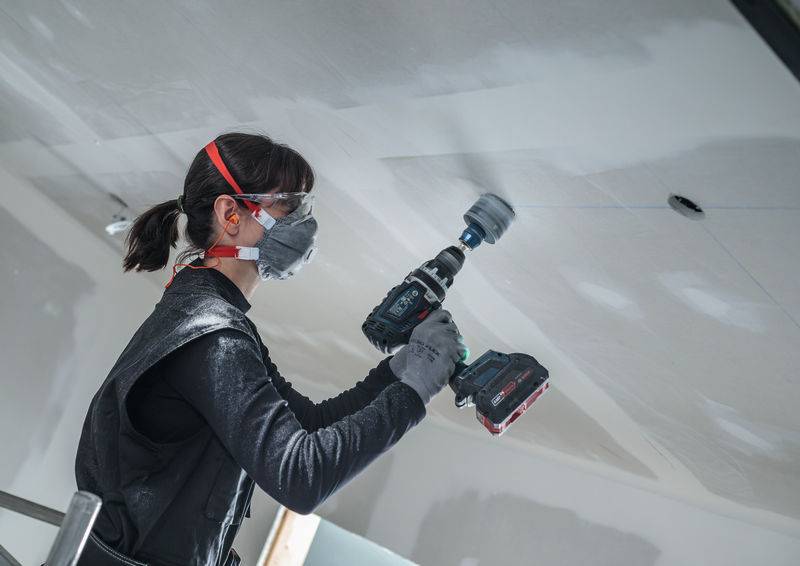 Eine Person in Schutzausrüstung nutzt einen Elektroschrauber, um eine kreisförmige Öffnung in einer Decke zu schaffen, was auf eine Bau- oder Renovierungsaufgabe hinweist.
