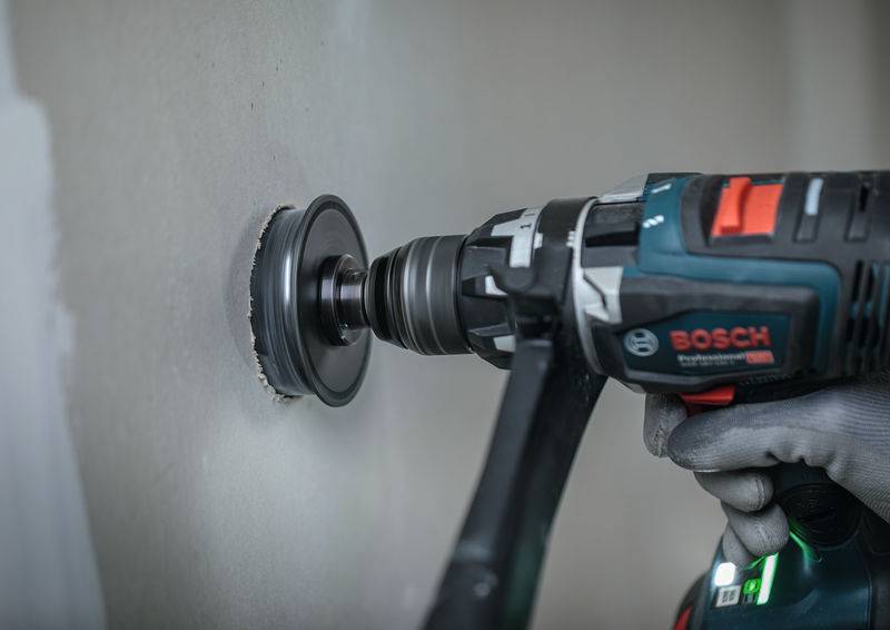 Eine Person benutzt eine Bosch-Bohrmaschine mit einem Lochsägen-Aufsatz, um ein kreisförmiges Loch in eine Wand zu schneiden, wobei sie graue Handschuhe trägt, um sicher zu arbeiten.