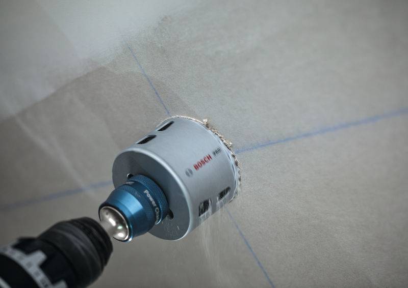 Ein Elektrobohrer mit einer kreisförmigen Schneidvorrichtung bohrt in eine Oberfläche und erzeugt dabei ein Loch. Um das Bohrwerkzeug sind feine Partikel sichtbar.