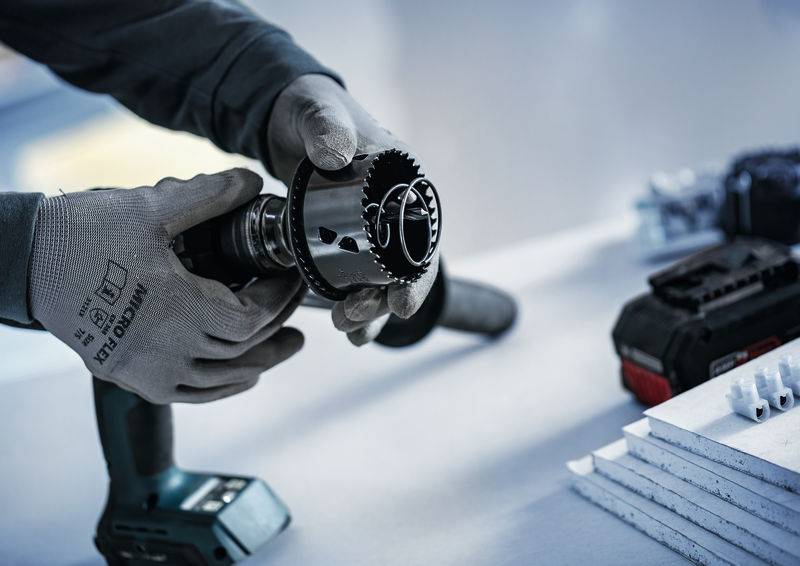 Eine Person mit Handschuhen hält einen Bohrer mit einer Lochsägenaufsatz und bereitet sich auf die Arbeit vor. Eine Batterie und weiße Materialien ruhen in der Nähe auf dem Tisch.