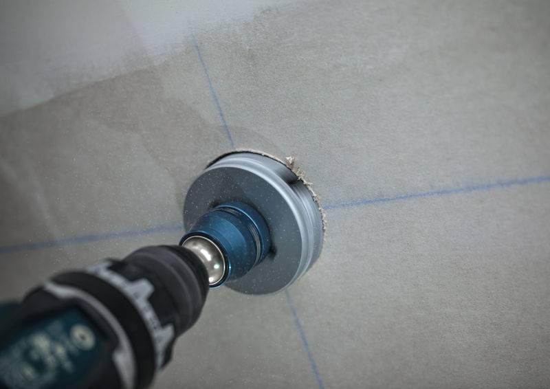 Ein Bohrer mit einem runden Aufsatz erstellt ein Loch in einer Fliese, das mit blauen Richtlinien gekennzeichnet ist und präzise Positionierung für die Installation zeigt.