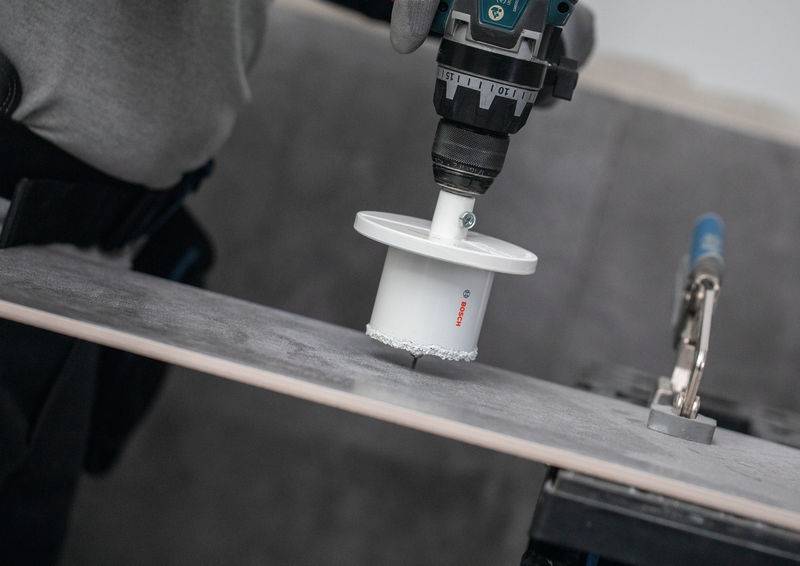 Eine Person benutzt einen Elektrobohrermaschine mit einer Lochsäge, um ein Loch durch eine Fliese zu schneiden, wahrscheinlich für Installations- oder Reparaturarbeiten.