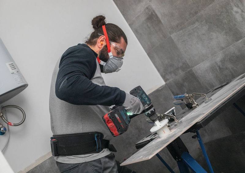Eine Person in Schutzausrüstung verwendet einen Elektroschrauber, um ein kreisförmiges Loch in eine Fliese auf einer Werkbank in einer Werkstattumgebung zu schneiden.