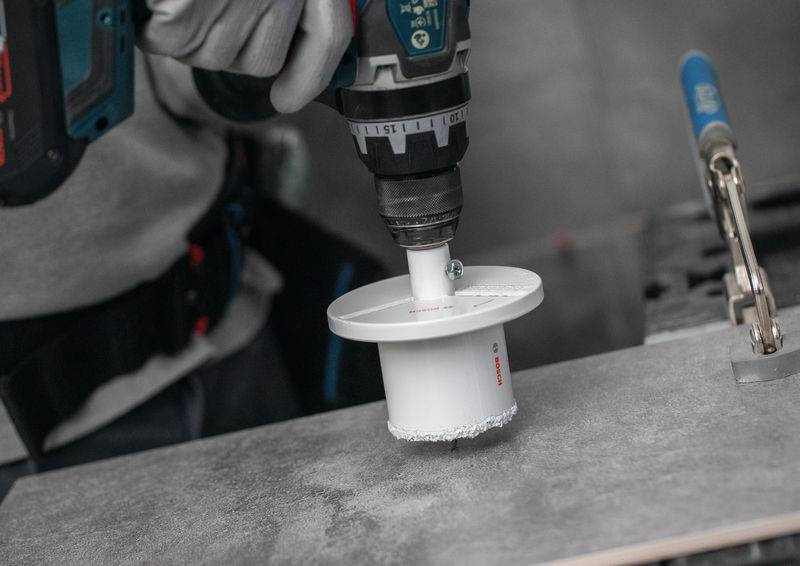 Ein Arbeiter verwendet eine Bohrmaschine mit einer Lochsäge, um ein kreisförmiges Loch in einer Fliese zu schneiden, was auf eine Bau- oder Renovierungsaufgabe hinweist.