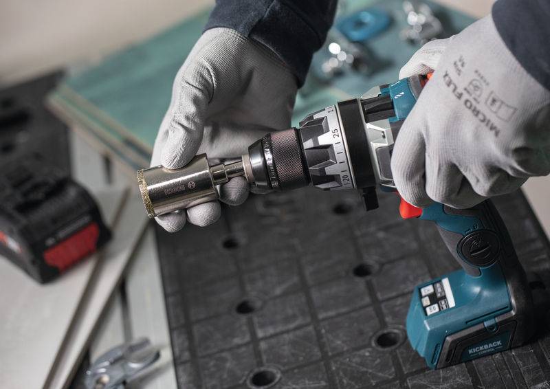 Eine Person mit Handschuhen benutzt einen kabellosen Elektroschrauber mit einer Steckaufsatzhalterung auf einer schwarzen Arbeitsfläche. Werkzeuge und Materialien befinden sich in der Nähe.