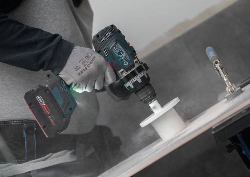 Eine Person verwendet eine Elektrobohrmaschine mit einer Lochsäge, um in eine ebene Oberfläche zu schneiden und erzeugt dabei Staub. Sie trägt Schutzhandschuhe und Arbeitskleidung.