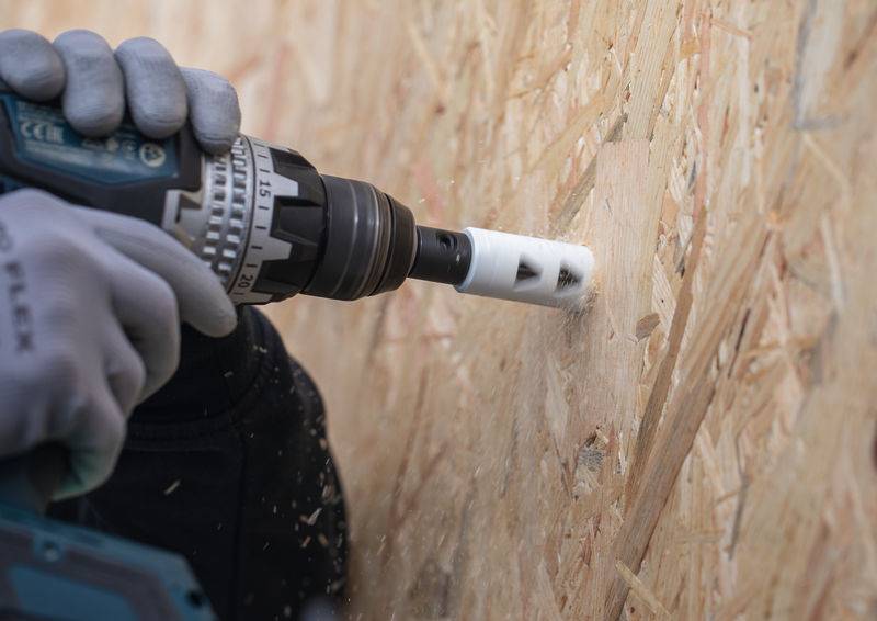 Eine Person mit Handschuhen benutzt eine Elektrobohrmaschine mit einem Lochsägenaufsatz, um ein Loch in ein Holzbrett zu bohren und konzentriert sich dabei auf den Bohrvorgang.