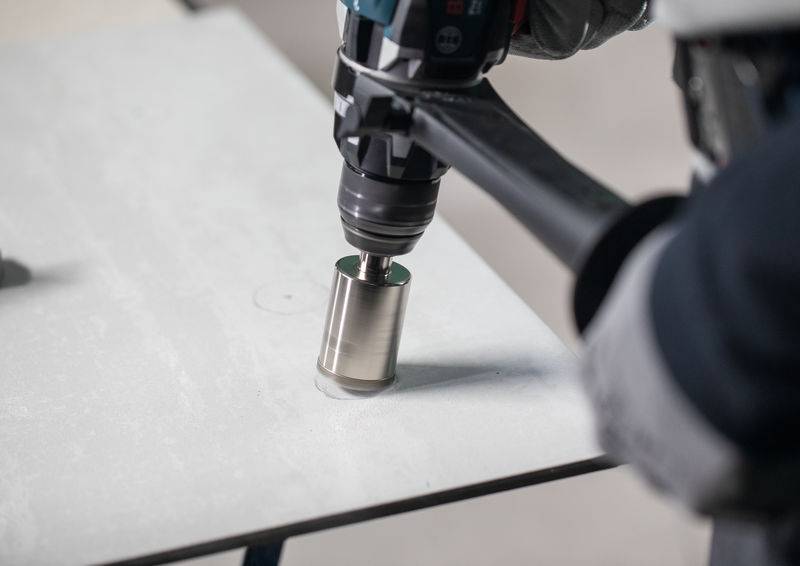 Eine Person benutzt eine Elektrobohrmaschine mit einem Lochsägen-Aufsatz, um präzise in ein Metallstück zu bohren und konzentriert sich dabei auf Genauigkeit und Technik.