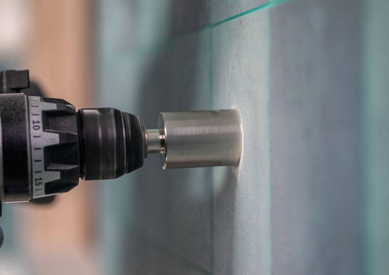 Eine Elektrobohrmaschine mit einer Lochsäge-Aufsatzbohrung durch eine Glasplatte, die einen Prozess zum Erstellen von kreisförmigen Löchern in harten Materialien anzeigt.