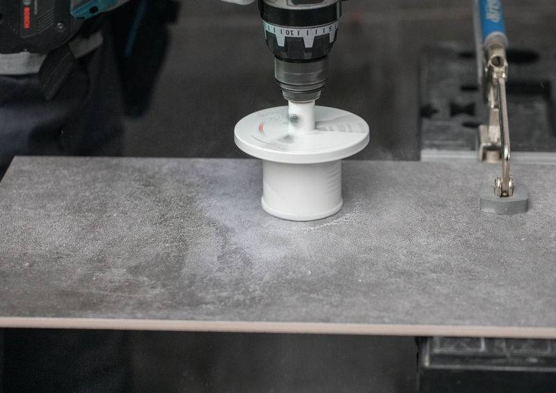 Ein Bohrer erzeugt einen kreisförmigen Schnitt in einer dunklen Fliese, während er in einem Schraubstock gesichert ist, und erzeugt dabei weißen Staub um die Bohrerstelle in einer Werkstattumgebung.