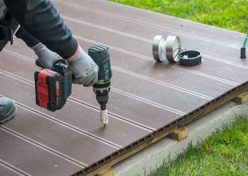 Person, die einen Elektroschrauber verwendet, um braune Terrassenbretter auf einem Holzrahmen zu befestigen, mit Werkzeugen und Gras im Hintergrund sichtbar.