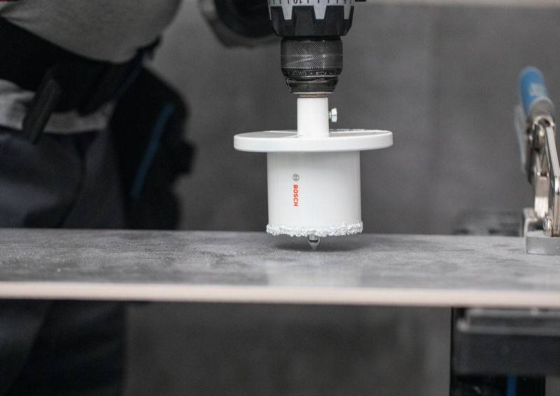 Eine Person benutzt eine Elektrobohrmaschine mit einem Lochsägen-Aufsatz, um durch eine ebene Oberfläche, möglicherweise Beton oder Fliesen, in einer Werkstattumgebung zu bohren.