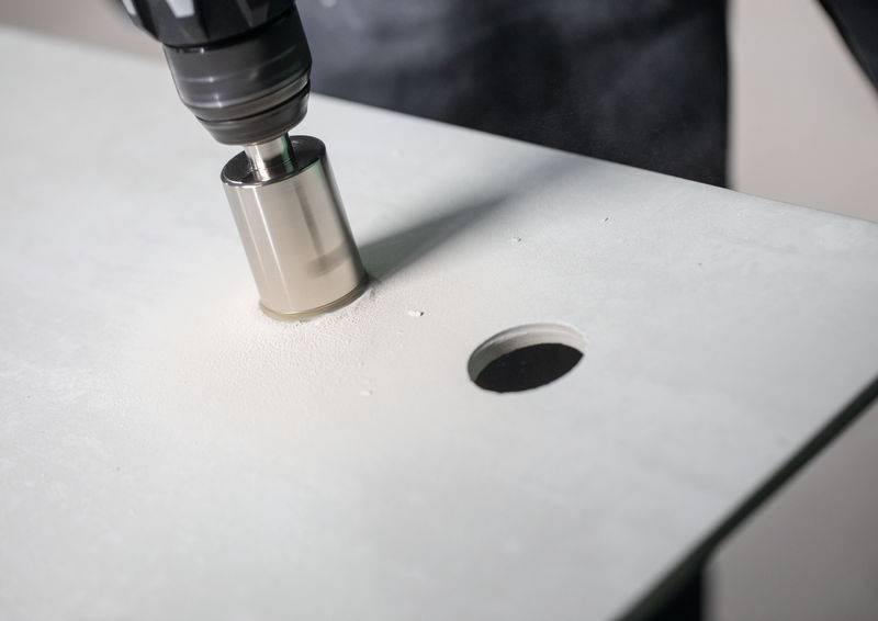 Ein Elektroschrauber mit einer Lochsägen-Aufsatzversion schneidet durch eine hellhäufige flache Oberfläche und erstellt ein rundes Loch, wobei Sägespäne sichtbar sind.