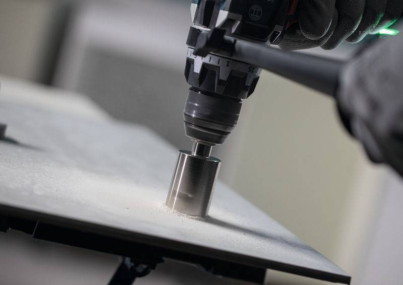 Eine Nahaufnahme einer behandschuhten Hand, die einen Elektrobohrer verwendet, um ein Loch in eine flache, metallische Oberfläche zu bohren, wobei Bauarbeiten hervorgehoben werden.