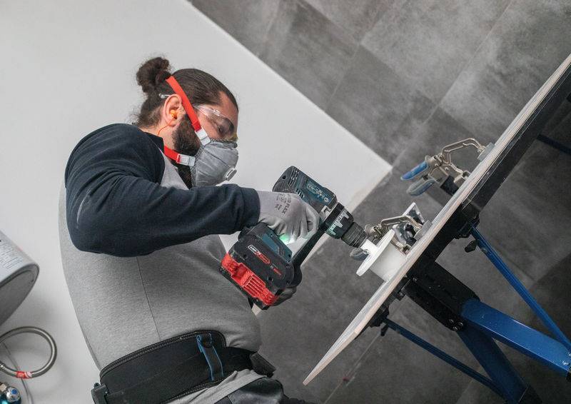 Eine Person benutzt einen Elektroschrauber, um mit Schutzbrille, Handschuhen und Maske für Sicherheit in einer Werkstatt ein Loch in eine Arbeitsfläche zu bohren.
