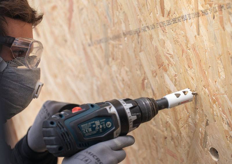 Eine Person, die eine Schutzbrille und eine Maske trägt, verwendet einen elektrischen Bohrer mit einem Lochsägenaufsatz, um ein Loch in eine Holzplatte aus OSB zu bohren.