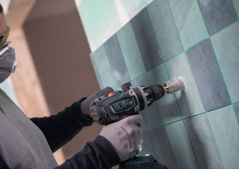 Eine Person trägt Handschuhe und eine Maske und verwendet einen Elektroschrauber, um Löcher in blaugrüne Keramikfliesen an einer Wand für die Installation zu bohren.