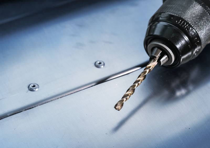 Eine Nahaufnahme einer Elektrobohrmaschine mit einem angebrachten Metallbohrer, positioniert auf einer metallischen Oberfläche mit zwei kleinen Schrauben. Die Szene deutet auf einen Bau- oder Reparaturkontext hin.