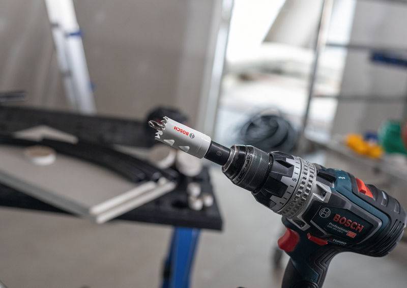 Ein Bosch-Elektrobohrer mit Lochsägen-Aufsatz ist im Vordergrund scharf fokussiert und vor einem unscharfen Werkstatt-Hintergrund mit Werkzeugen positioniert.