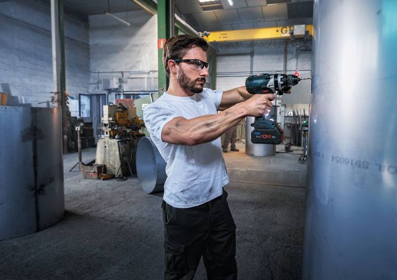 Mann mit Schutzbrille benutzt einen Elektroschrauber auf Metall in einer Werkstatt.